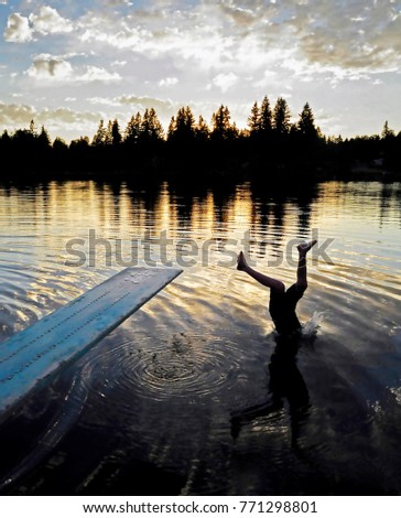 dark silhouette of Boy upside down mid dive and half his body submerged under the water with reflection in the water in a warm evening light with dark silhouette of trees on the horizon