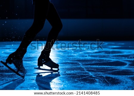 Dark silhouette of female legs in figure skating skates sliding on ice arena. Young woman trains on dark ice rink with blue light. Shiny smooth surface of ice with scratches from skates. Close up.