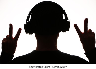 Dark silhouette in headphones listens to music on a white background. A man in headphones listens to music and holds two fingers in the upward sign of victory.