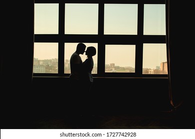 Dark Silhouette Of A Loving Couple, Man And Woman No Faces, Against Of A Huge Stained Glass Window With A View Of The City And Curtains On The Sides. The Comfort Of Home, A Life Together For Couples