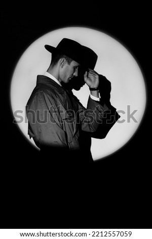A dark silhouette of a male detective in a coat and hat in the noir style. A dramatic portrait in the style of detective films of the 1950s. A silhouette in a circle of light, like Agent 007.