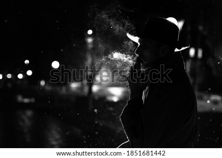 dark silhouette of a man in a hat Smoking a cigarette in the rain on a night street in the style of Noir