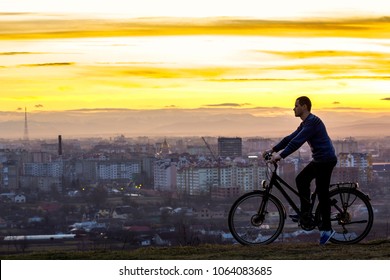 Dark silhouette of man standing near a sports bicycle with the night city view behind. Active life and travel concept. Sunset over Ivano Frankivsk city in Ukraine with lonely cyclist enjoying view.