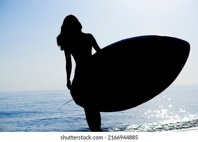 The dark silhouette of a slender girl surfer walks along the ocean and holds a board for  SUP water sports in the rays of the sunset. - Shutterstock ID 2166944885