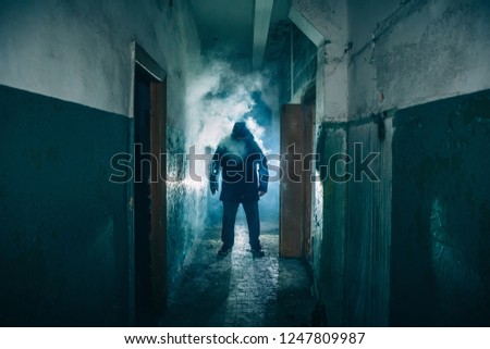 Dark silhouette of strange danger man in hood in back light with smoke or fog in scary grunge corridor or tunnel, toned