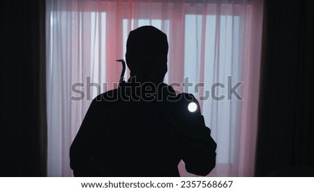 The dark silhouette of a thief in the evening in an apartment illuminated by red light from police sirens. Robber in a black balaclava, with a flashlight and with a crowbar.