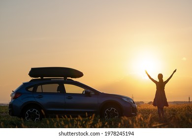 Dark Silhouette Of Woman Driver Standing Near Her Car On Grassy Field Enjoying View Of Bright Sunset. Young Female Relaxing During Road Trip Beside SUV Vehicle.