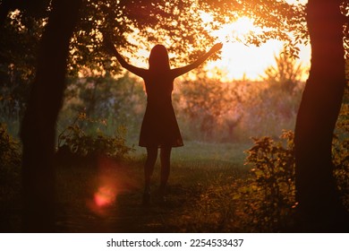 Dark silhouette of young woman walking alone through dark forest joyfully raising her hands in summer evening. Enjoying nature and outdoor activities concept - Shutterstock ID 2254533437