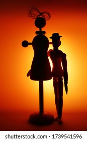 Dark Silhouettes Of Two Wooden Dolls In The Form Of A Mannequin And Gestalts In A Contoured Orange Light. Close Up. A Funny Picture On The Topic Of Love Relationships Between Very Different People.