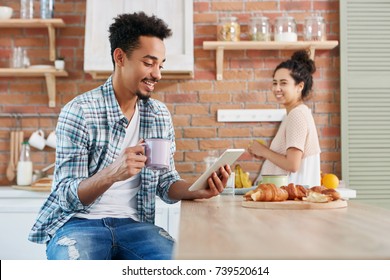 Dark skinned bearded hipster guy makes video call over tablet computer, holds mug of coffee, eats tasty croissants and his wife stands in background, prepares fruit salad, tries to eavesdrope talk - Powered by Shutterstock