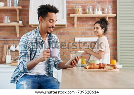 Dark skinned bearded hipster guy makes video call over tablet computer, holds mug of coffee, eats tasty croissants and his wife stands in background, prepares fruit salad, tries to eavesdrope talk