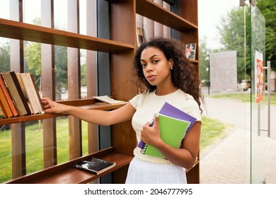 Dark Skinned Brunette Girl In Library. Mixed Race Female With Books And Notebooks Campus. Portrait Cheerful African American Young Woman With Loose Wavy Curls Black Hair.