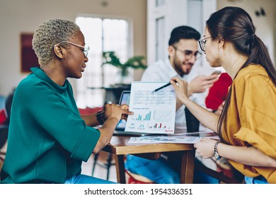 Dark skinned and Caucaisan female partners analyzing graphic charts during paperwork briefing at table desktop, young diverse colleagues talking about infographics statistics while cooperate on report - Powered by Shutterstock