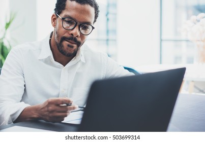 Dark Skinned Coworker Wearing Glasses And Using Laptop In Modern Office.African American Man In White Shirt Working On Workplace.Horizontal,blurred Background.