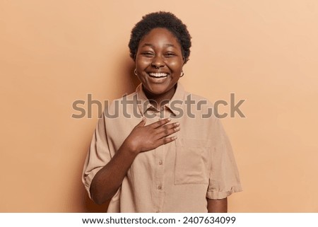 Dark skinned curly woman with toothy smile keeps hand on chest expresses sincere emotions being in good mood wears shirt and piercing in nose isolated on brown background. People and happiness concept