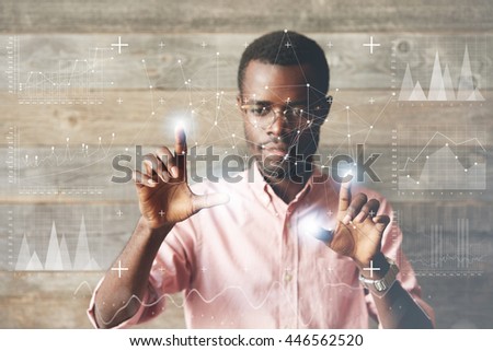 Dark skinned entrepreneur working with virtual screen, navigating and changing positions of icons on futuristic technology display with fingers, looking at the panel absorbedly with excitement