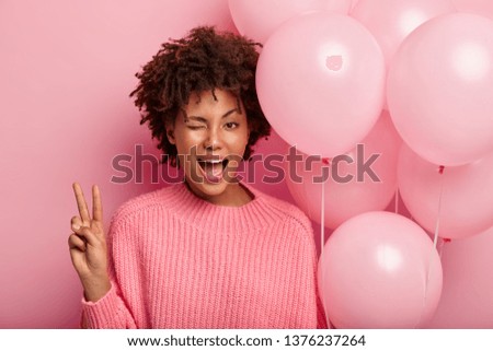 Dark skinned female model blinks eye, shows peace gesture, wears pink jumper and carries air balloons, has fun in circle of best friends, isolated against rosy background. Youth, celebration concept