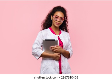 Dark Skinned Female Physician Medical Advisor Holding Digital Tablet With Medical History In Electronic Clinical Record On Pink Studio Wall. Woman Medical Student, Intern, GP In Eyewear And White Coat