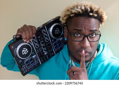 Dark Skinned Guy With A Serious Expression In A Blue Hoodie Holds A DJ Remote On His Shoulder. Young Black Man In Glasses With An Afro Hairstyle Presses His Finger To His Mouth, Calling For Silence.