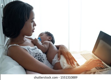 Dark Skinned Mother Holding Newborn Baby While Sitting On Bed With Laptop And Checking Mail With Stress, Tired Dark Skin Working Mom With Sleeping Child In Her Lap