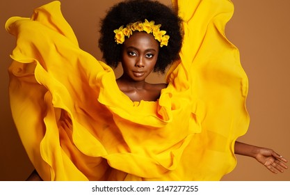 Dark Skinned Woman Fashion Portrait In Yellow Flying Dress. Beauty African Model With Flower Wreath In Black Afro Hairstyle. Dancer With Fluttering Silk Fabric Over Studio Beige Background