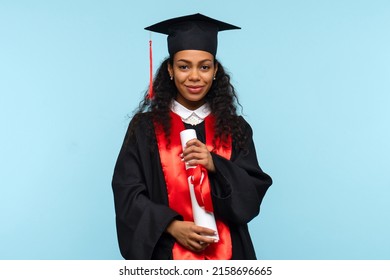 Dark Skinned Woman Graduate Wearing Ceremony Robe And Graduation Cap Holding Certificate On Blue Background. Girl Celebrating Graduation And Getting Diploma. Degree Paper. Education Concept