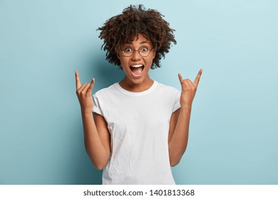 Dark skinned woman shouts from happiness, has widely opened mouth, makes rock n roll gesture, wears casual outfit, says rock this party, feels cool, models over blue background. Body language - Shutterstock ID 1401813368