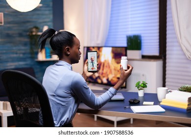 Dark Skinned Woman Showing Pills Bottle To Doctor During Video Call Late At Night From Living Room. Black Patient In A Video Call With Medic Discussing Health Problems Of Woman.