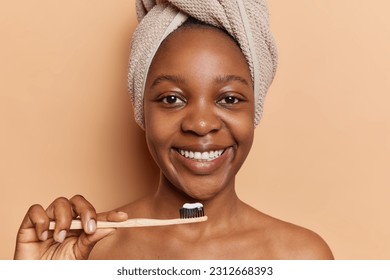 Dark skinned young woman stands against brown background uses toothbrush toothpaste for her oral hygiene diligently cares for teeth stands shirtless has bath towel on head isolated on brown background - Powered by Shutterstock