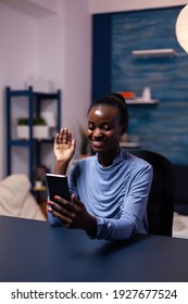 Dark Skinnned Woman Saying Hello In The Course Of Video Conference Sitting At Desk Late At Night In Office. Black Freelancer Working With Remotely Team Chatting Virtual Online Conference.