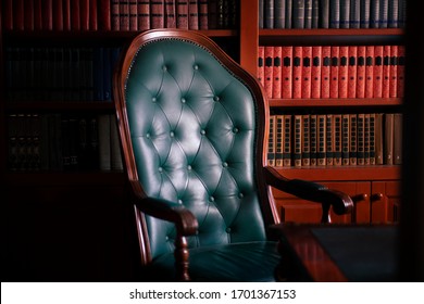 Dark solid interior of old library with many books on the wooden shelves. Vintage classic chair from green leather and red wood, antique table, luxury design. Indoors, copy space.