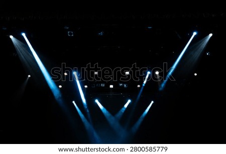 Dark stage featuring multiple blue spotlights casting sharp beams through theatrical haze.