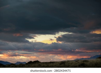 Dark storm clouds gather over a glowing sunrise horizon above windswept grassland and distant mountain peaks in Patagonia.