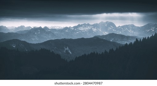 Dark storm clouds hovering over a panoramic view of layered mountains with snow capped peaks and evergreen forests in winter