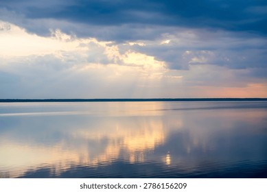 Dark storm clouds and soft sunset light above peaceful lake with reflections on smooth water surface