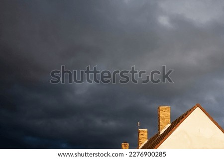 Dark stormy clouds in the background,bottom right corner of image shows the roff of a detached house with three orange brick chimneys sticking up .