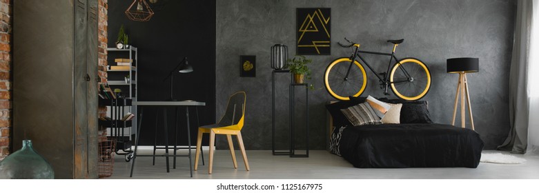 Dark studio flat interior with textured grey wall, black and yellow bike standing on bedhead, desk with geometric chair and double bed with black bedding