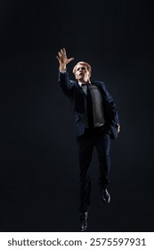 In a dark studio, a man in a sharp navy suit leaps mid-air, reaching upwards with determination. dynamic pose highlight ambition and success.