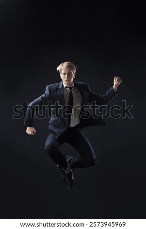 In a dark studio, a man in a sharp navy suit leaps mid-air, reaching upwards with determination. dynamic pose highlight ambition and success.