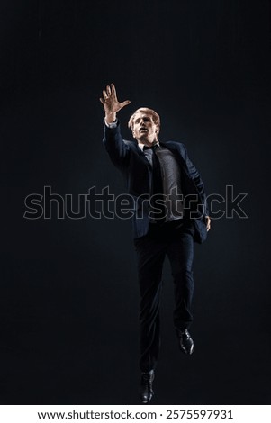 In a dark studio, a man in a sharp navy suit leaps mid-air, reaching upwards with determination. dynamic pose highlight ambition and success.