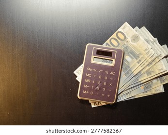 Dark surface holds calculator and stack of bills. Calculator rests atop fanned currency notes. Brown casing contrasts with bright paper bills. Numbers and symbols clearly visible on display