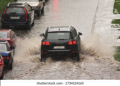 A dark SUV car drives fast with splash water on big puddle down the street next to parked vehicles in heavy rain in the city on a spring day, urban off-road back view - Shutterstock ID 1734524165