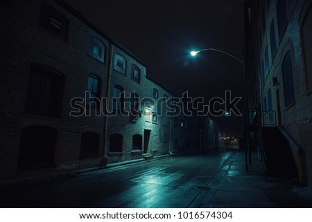 Dark urban city alley at night after a rain featuring vintage warehouses.