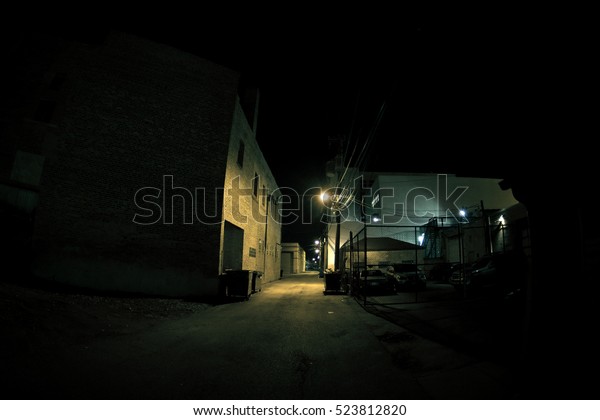 Dark urban city alley at night with fence, cars and\
parking lot