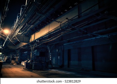 Dark Urban Downtown City Alley At Night With Garbage Trash Bins, Car Garage Doors And Elevated Train Tracks