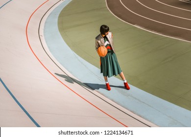 Dark-haired Appealing Girl. Tall Skinny Woman Standing With Orange Ball On The Stadium In Weird Outfit