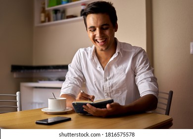 Dark-haired boy making notes in a notebook and drinking coffee. Teenager drawing and drinking something at wooden table. Man thinking and writing while using his white mug.