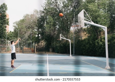 Dark-haired Boy Shooting A Street Basketball Free Throw. Image From Behind A Fence Of A Lone Young Man Scoring A Two-point Basket On A Blue Court. Friendship And Sport Concept