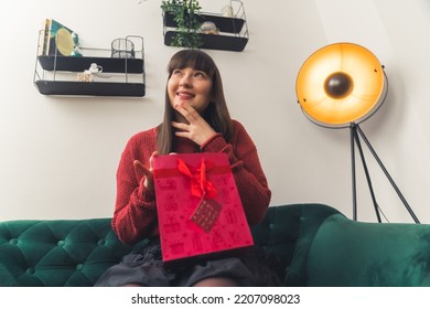 Dark-haired Caucasian Woman Wearing Red Sweater Opening Gift In Red Bag Looking Up Sitting On Green Sofa. Indoor Shot. High Quality Photo