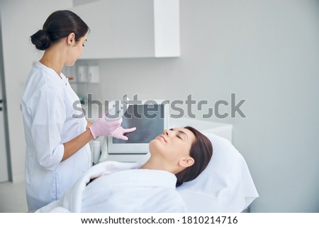 Dark-haired doctor in a lab coat and latex gloves touching the beauty machine monitor screen